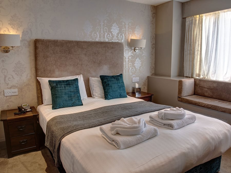 A neatly made bed with a headboard, two bedside lamps, a bedside table with a phone, and a window with curtains.