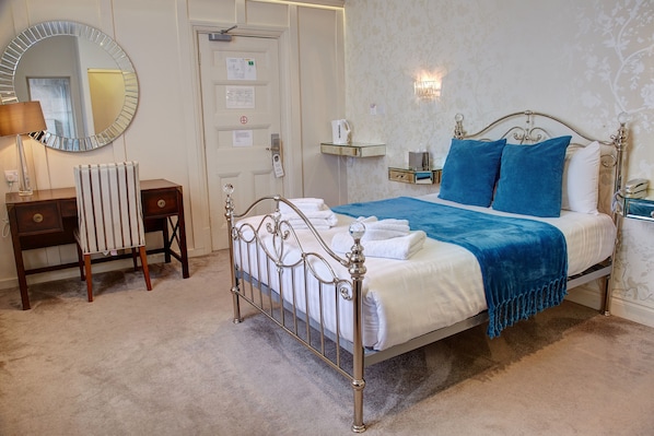 A neatly arranged bedroom with a metal-framed bed, a bedside table with a lamp, a mirror, and a radiator. - Orwell Hotel (Felixstowe)