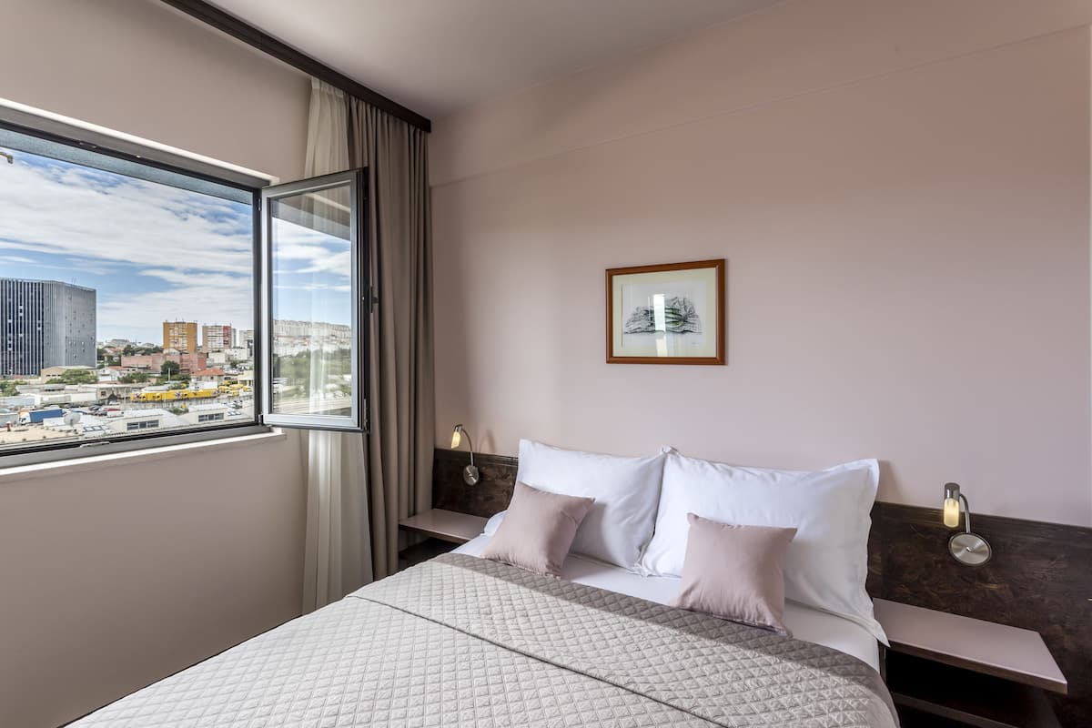 Habitación de hotel con una cama grande, una mesita de noche, un cuadro en la pared y una ventana con vistas a la ciudad.