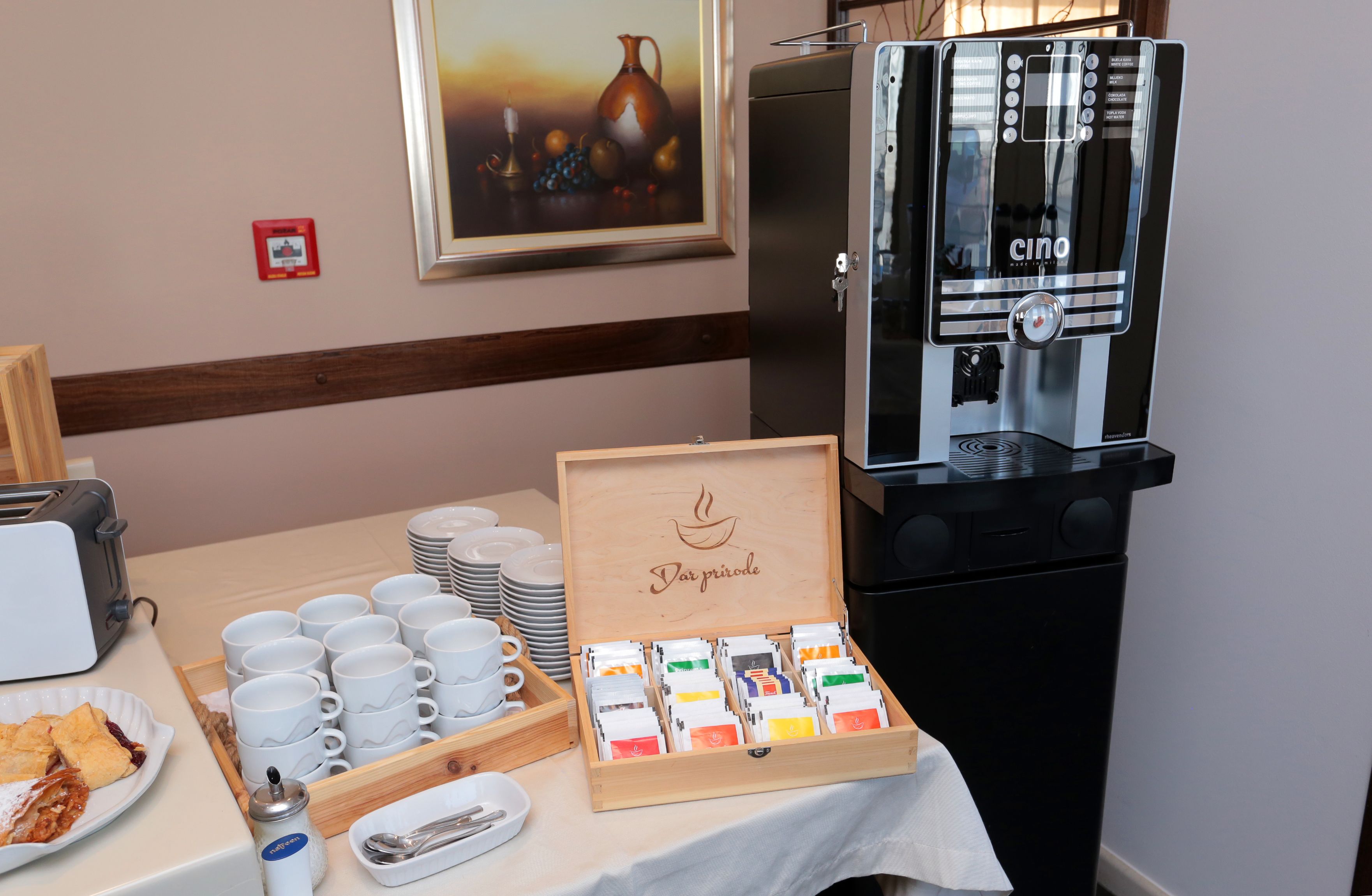 A breakfast setup with a coffee machine, tea and coffee packets, and a wooden box labeled "Dar priode".