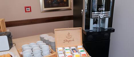 A breakfast setup with a coffee machine, tea and coffee packets, and a wooden box labeled "Dar priode".