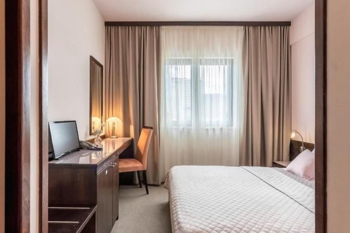 Une chambre d’hôtel avec un lit, un bureau avec un ordinateur portable, une chaise et une fenêtre avec des rideaux.