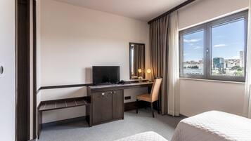 Habitación de hotel con escritorio, una silla, cama, televisor, ventana con vista a la ciudad y espejo.