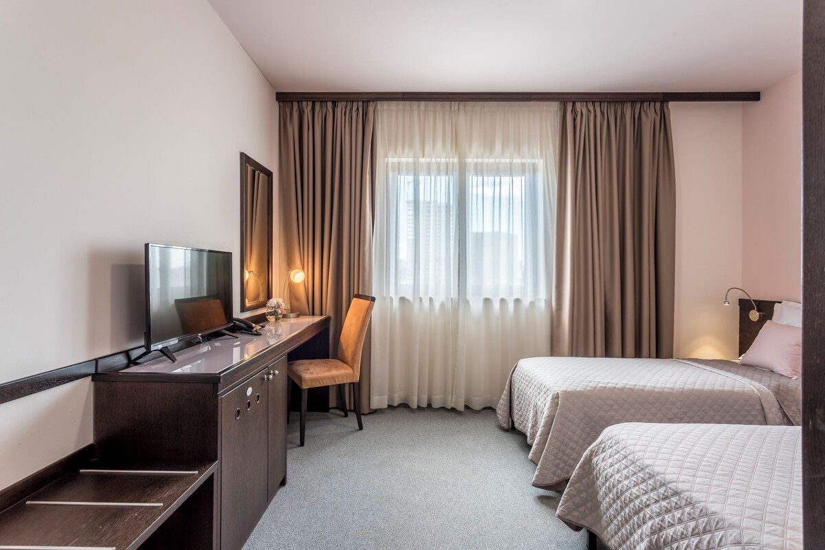 A hotel room with a desk, chair, two beds, a TV, and a window with curtains.