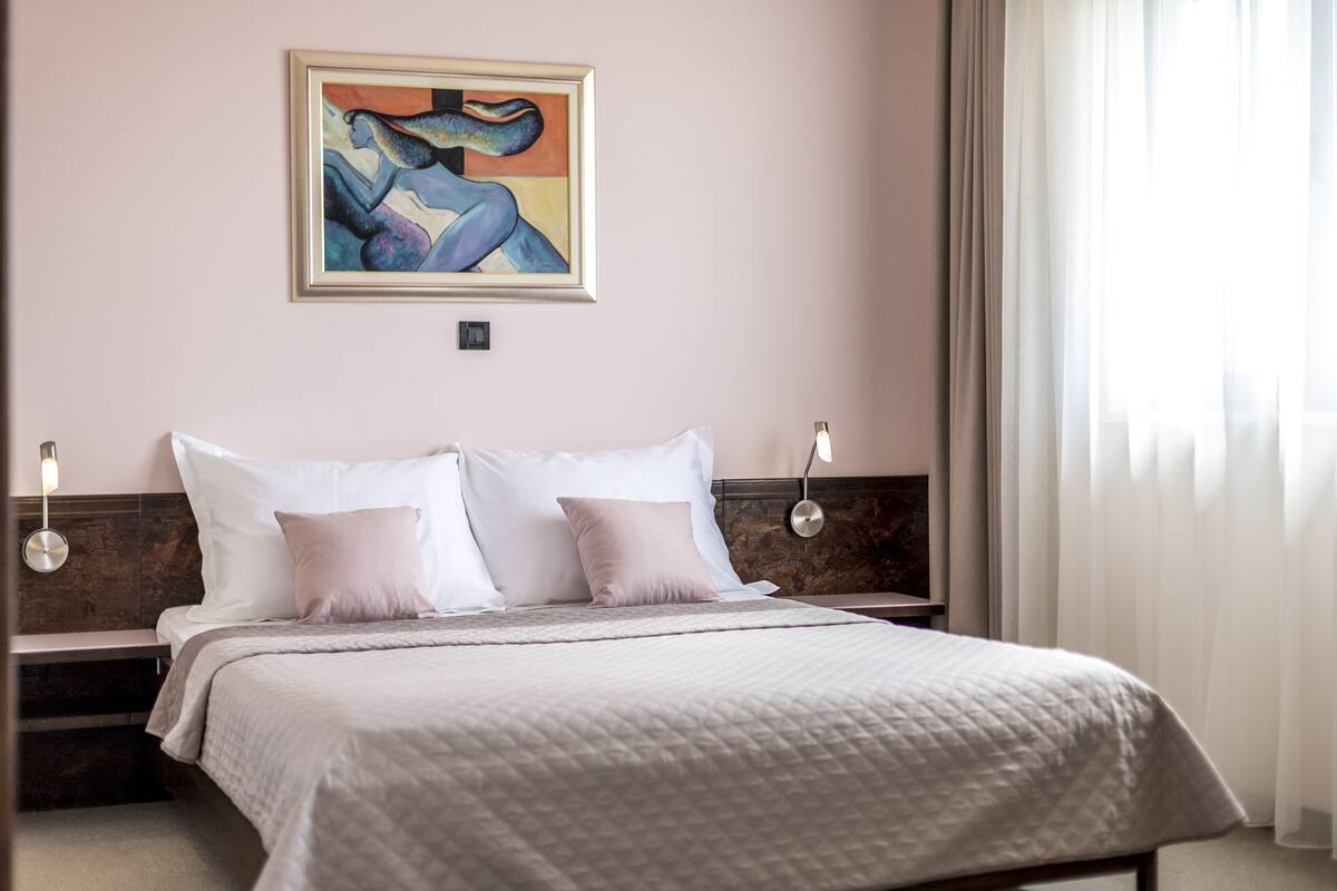 Una habitación de hotel con cama, un cuadro en la pared y una ventana con cortinas.
