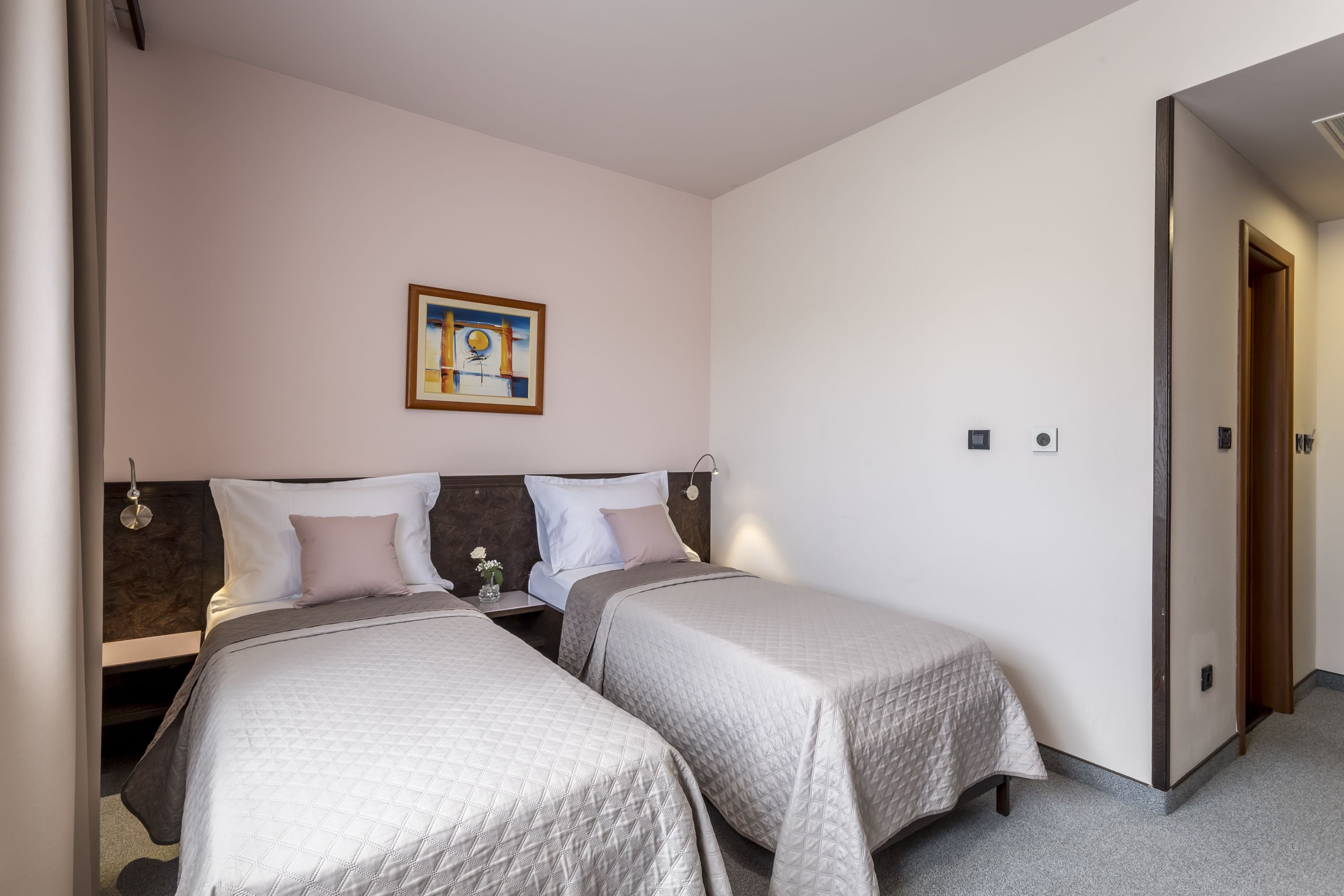 Une chambre d’hôtel avec deux lits, une tête de lit, un tableau encadré au mur et une porte donnant sur une autre pièce.