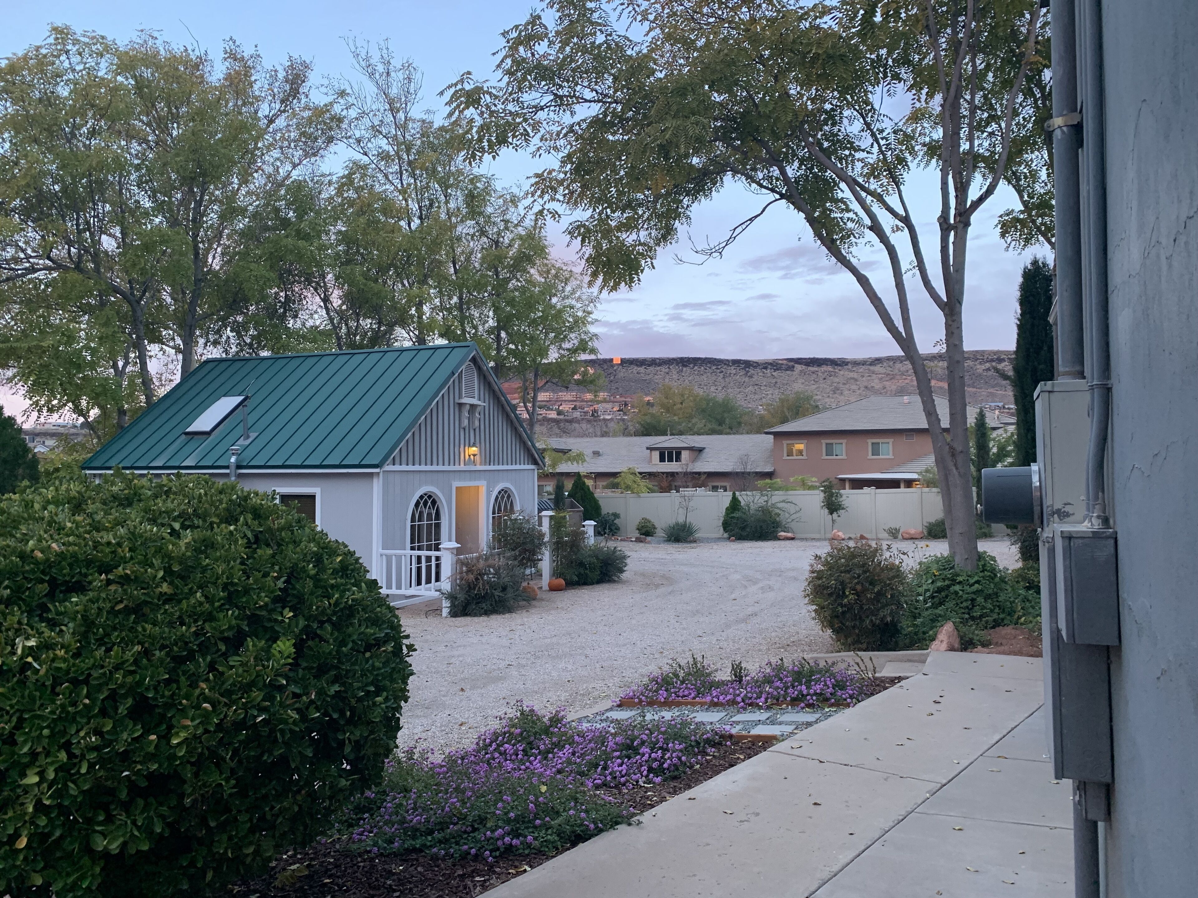 A residential area with a house featuring a green roof, a gravel driveway, and a flower bed with purple flowers.