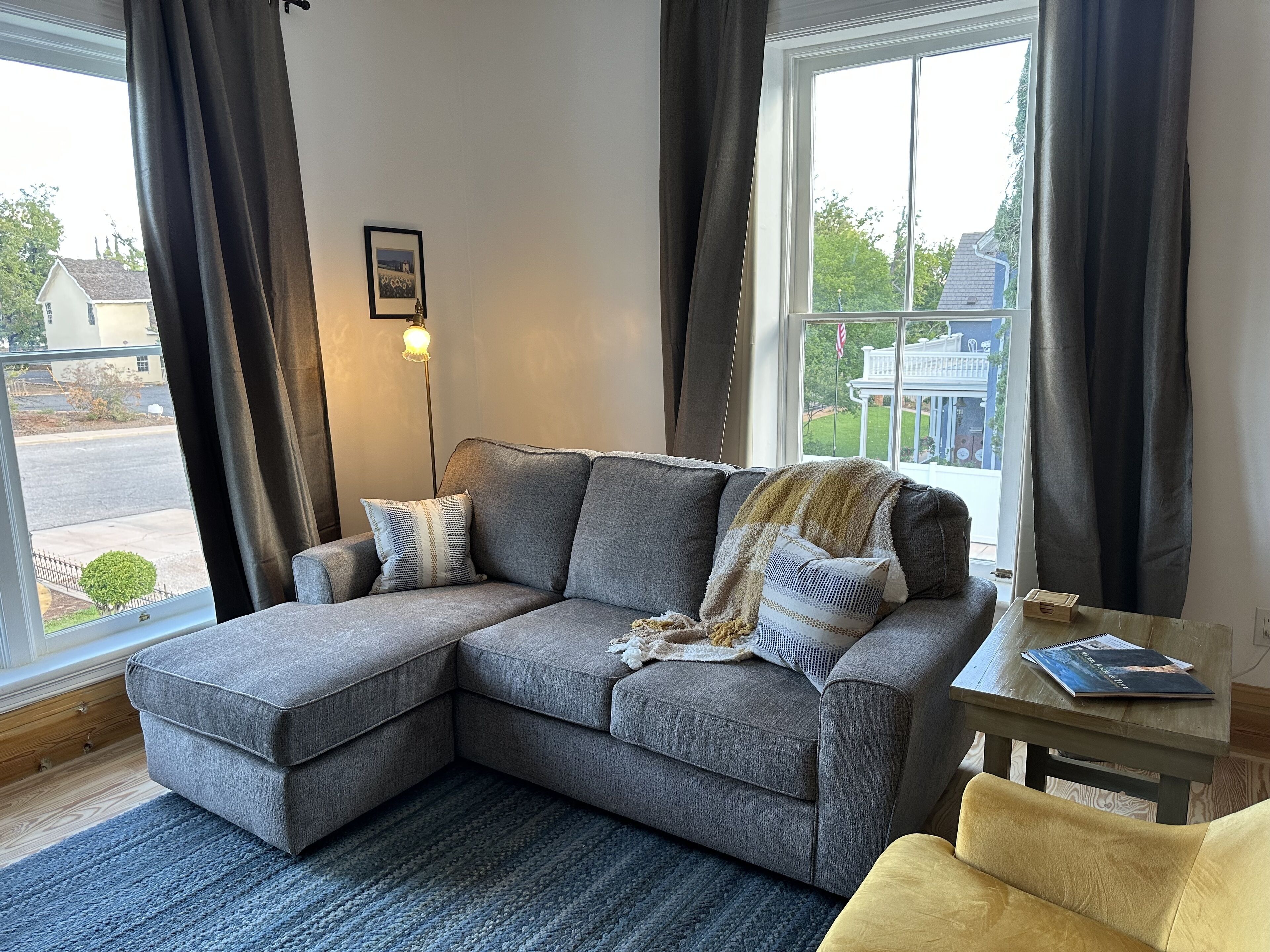 A gray sectional sofa with a throw blanket and pillows, a small wooden table with a book, and a view of a residential area through large windows.