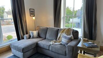 A gray sectional sofa with a throw blanket and pillows, a small wooden table with a book, and a view of a residential area through large windows.