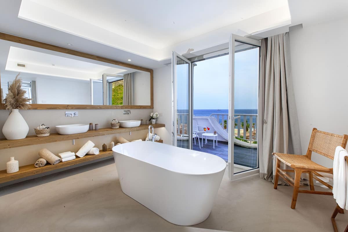 Une salle de bain moderne avec une baignoire sur pieds, un grand miroir et une vue sur la mer grâce à une porte en verre.