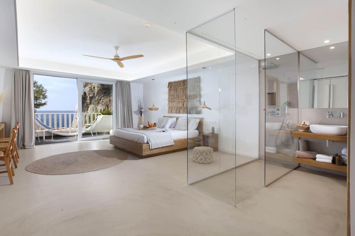 Une chambre moderne avec un grand lit, une cloison en verre, une salle de bain avec un lavabo et un miroir, et un balcon avec vue sur la mer.