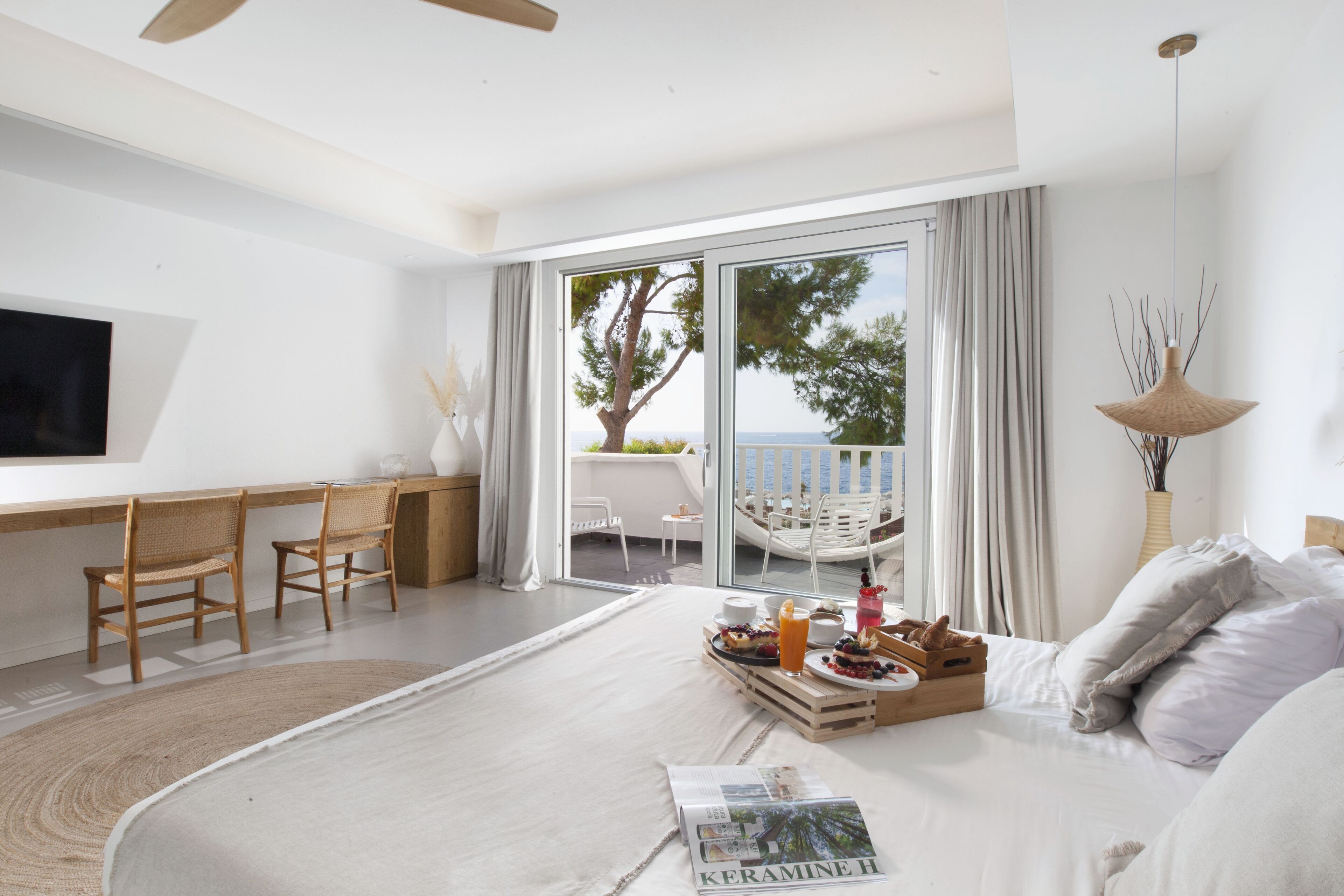 Quarto de hotel moderno com uma cama grande, área de jantar com cadeiras, televisão e vista para o mar através das portas da varanda.