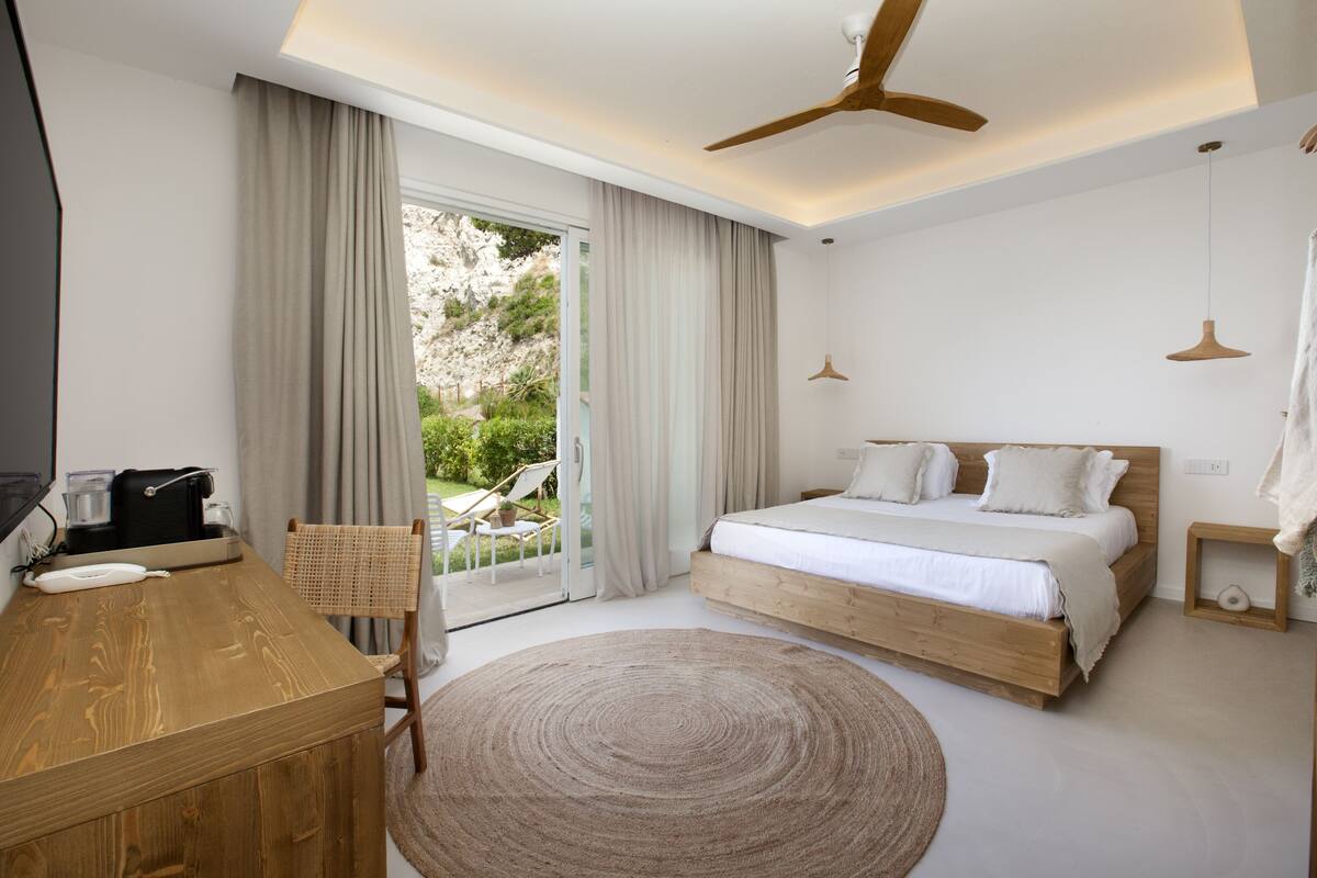 Une chambre moderne avec un lit en bois, un bureau et une chaise. On y trouve une grande fenêtre avec des rideaux, un ventilateur de plafond et un tapis rond.