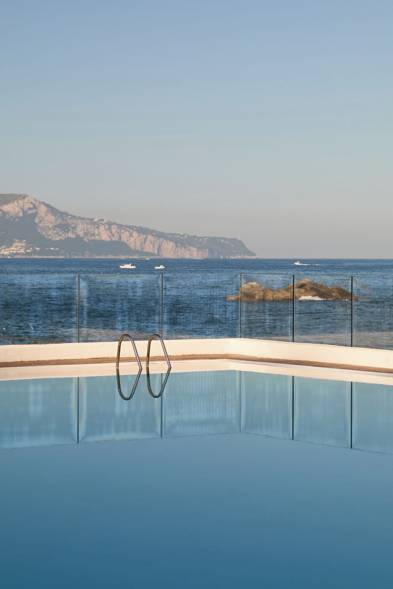 A clear blue pool with a metal ladder, surrounded by a glass railing, overlooking a calm sea with distant rocky cliffs.