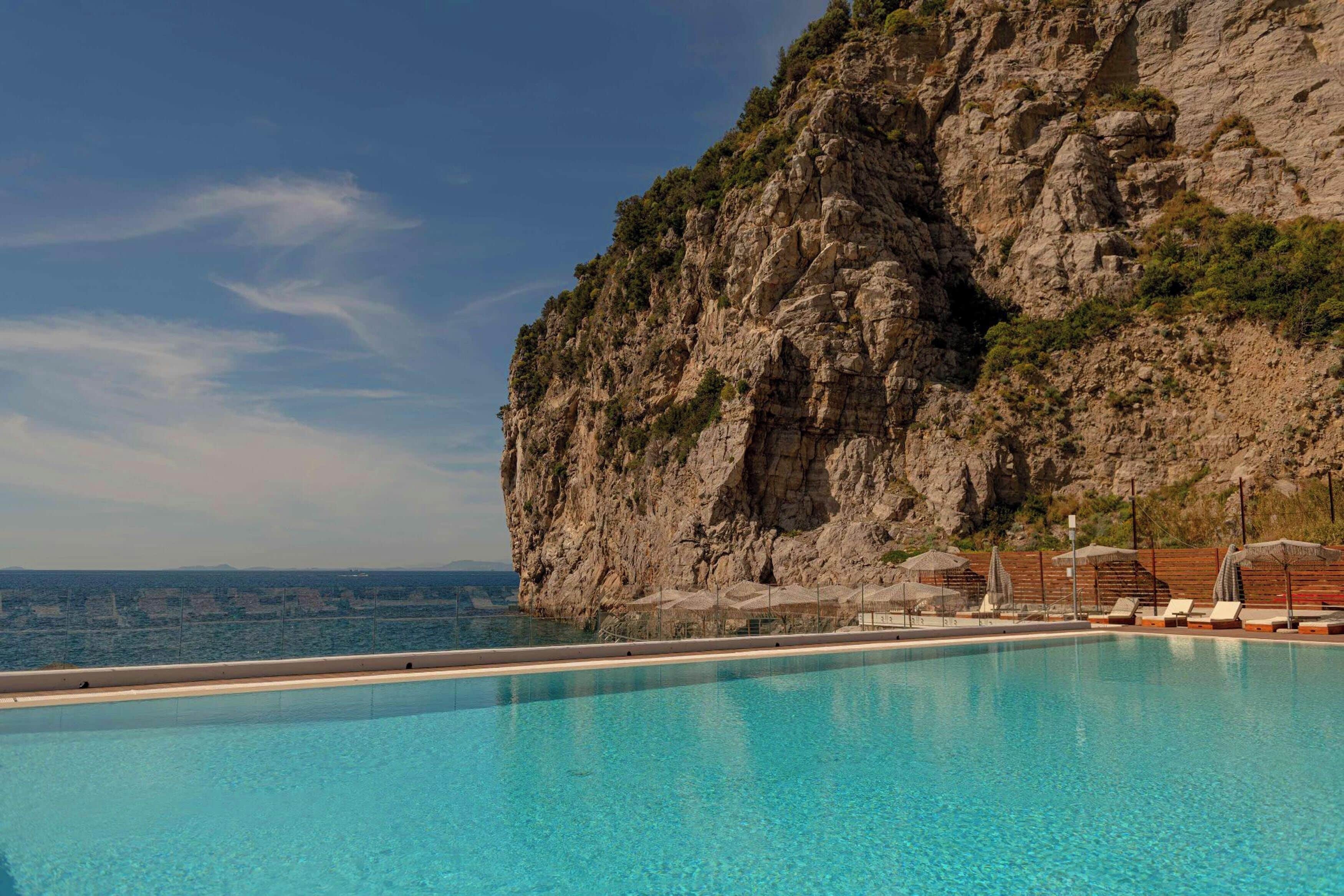 Una piscina azul cristalina con sillones de descanso y sombrillas, un acantilado rocoso y vistas al mar.
