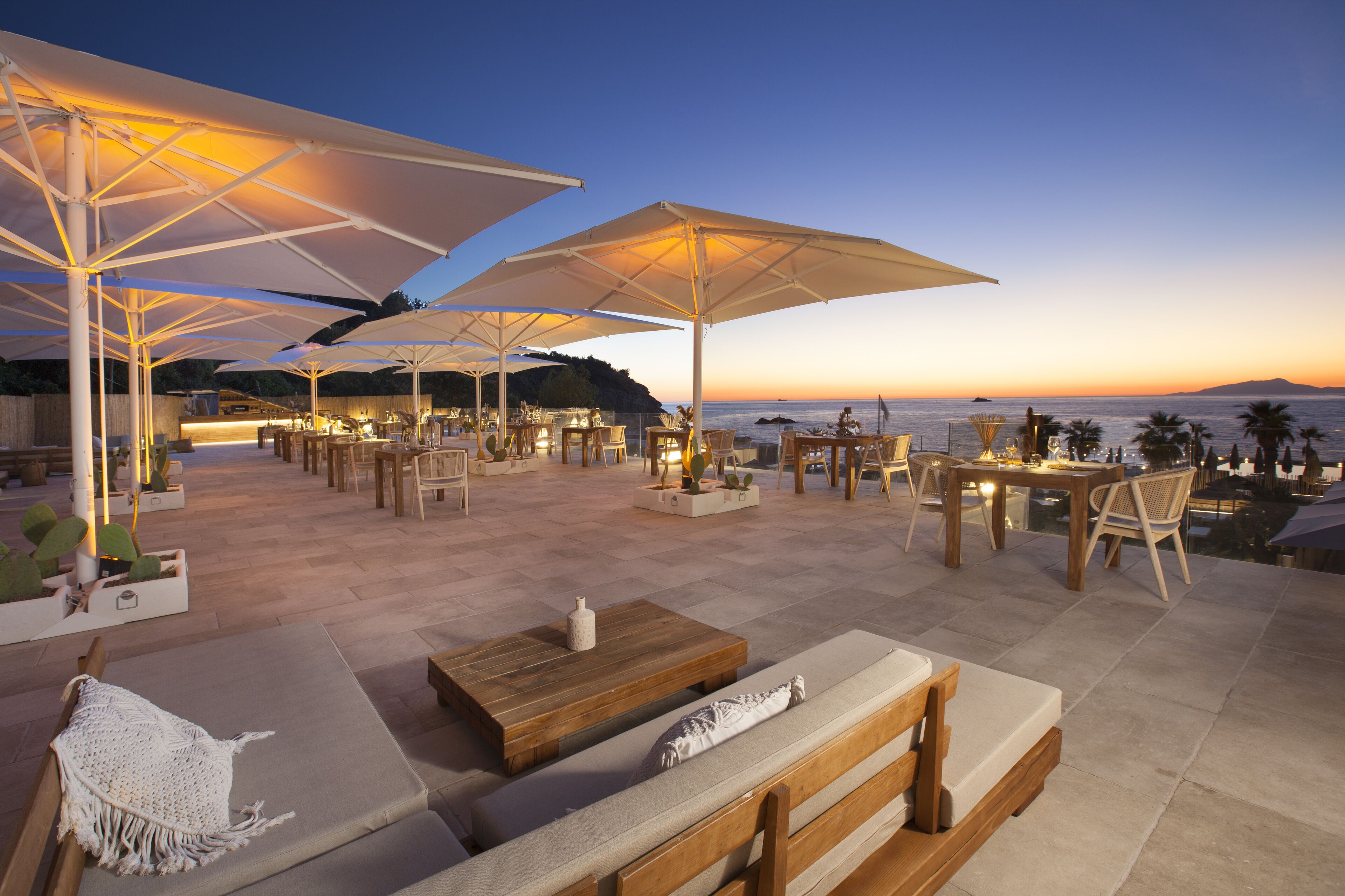 Un espace extérieur aménagé avec des tables et des chaises, des parasols, et une vue sur la mer au coucher du soleil.