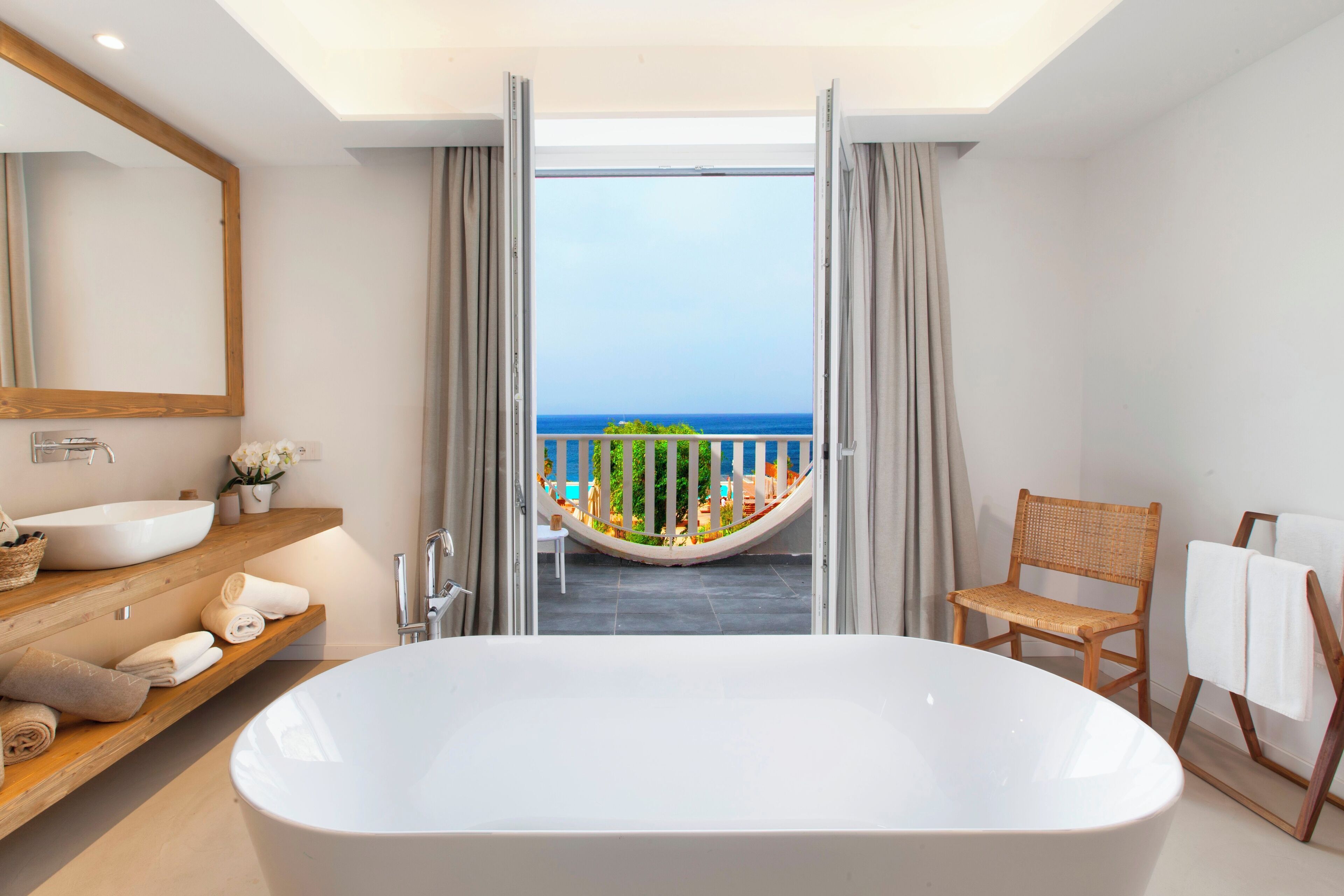 Une salle de bain moderne avec une grande baignoire, un meuble-lavabo en bois et une vue sur la mer par une porte ouverte.