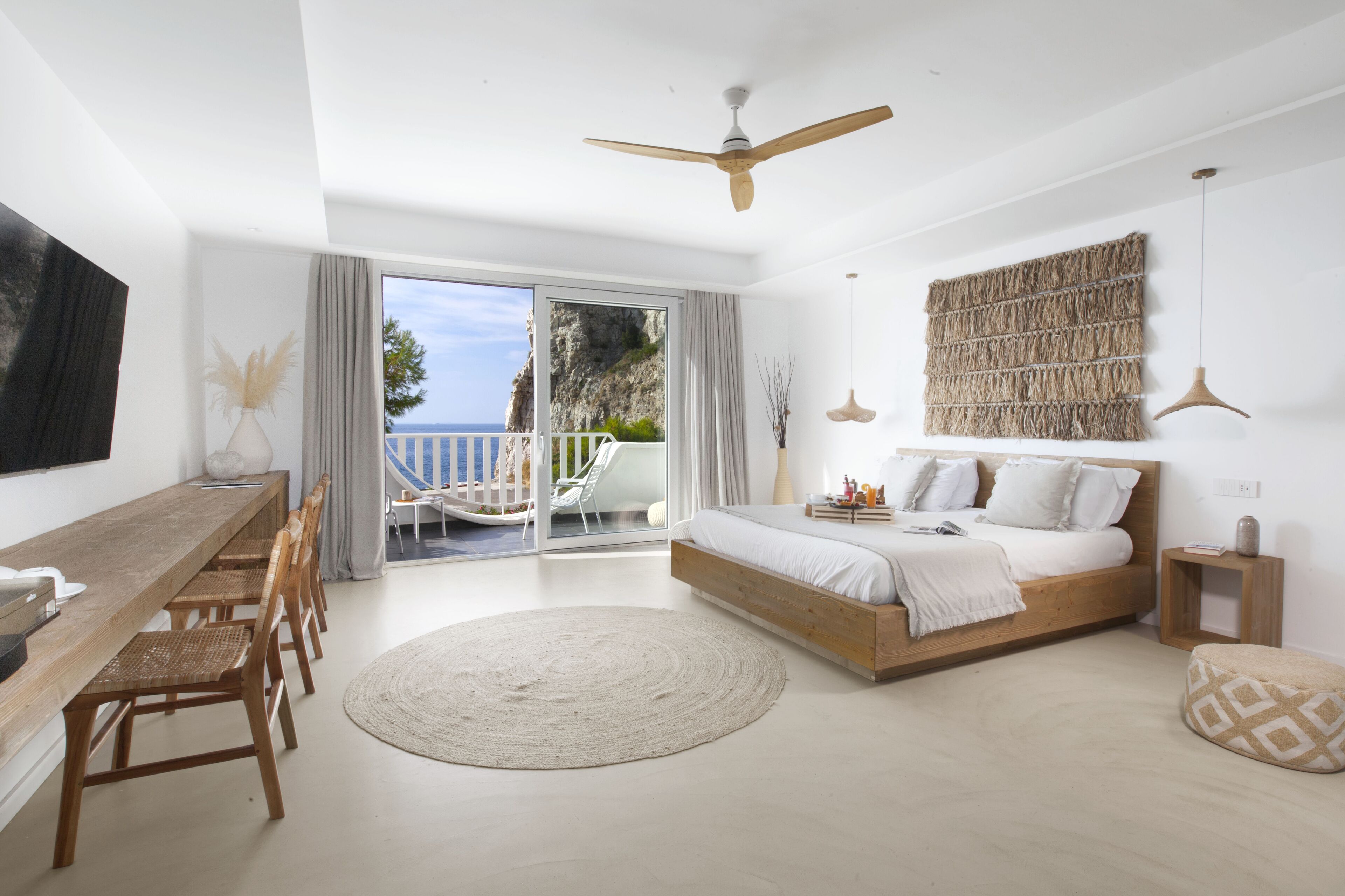 Quarto com cama, escrivaninha de madeira, cadeiras, tapete redondo, ventilador de teto e vista para o exterior.