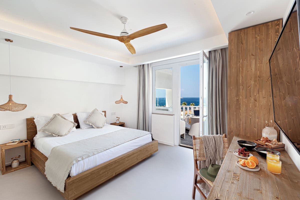 Une chambre d’hôtel moderne avec un grand lit, un ventilateur de plafond en bois, un balcon avec vue et une table préparée pour le petit-déjeuner.