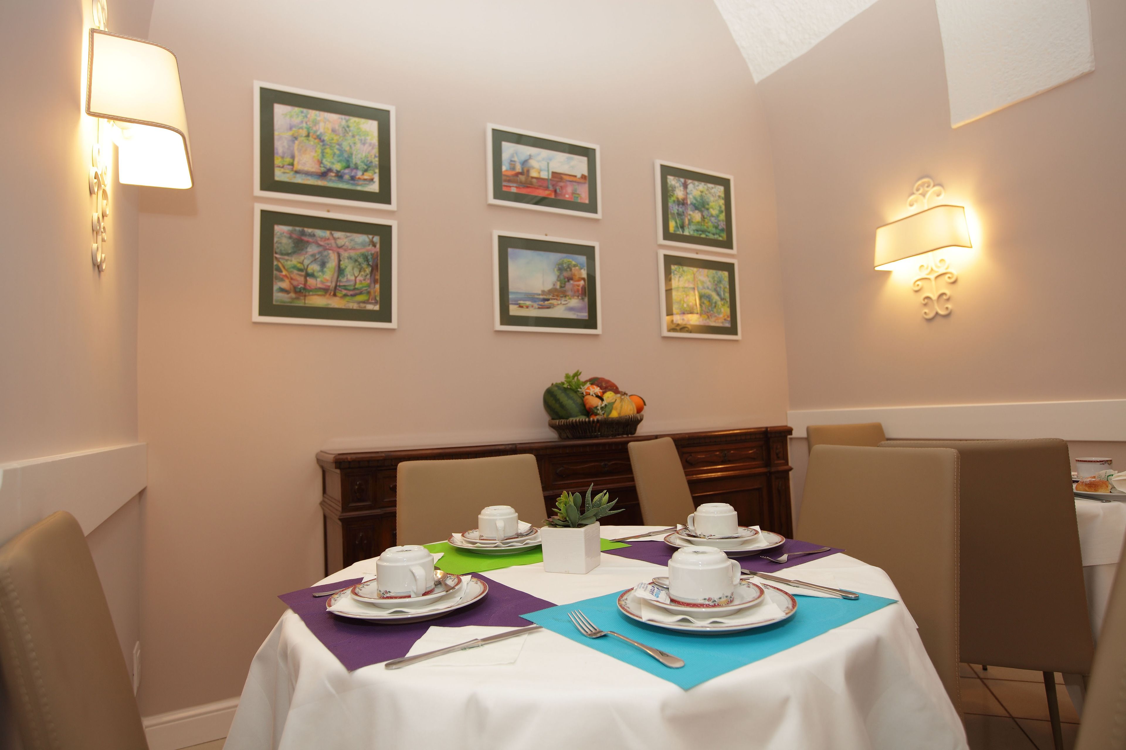A dining area with tables set for four, each with white tablecloths, purple and blue placemats, and white tableware. There are framed artworks on the wall and a fruit bowl on the table.