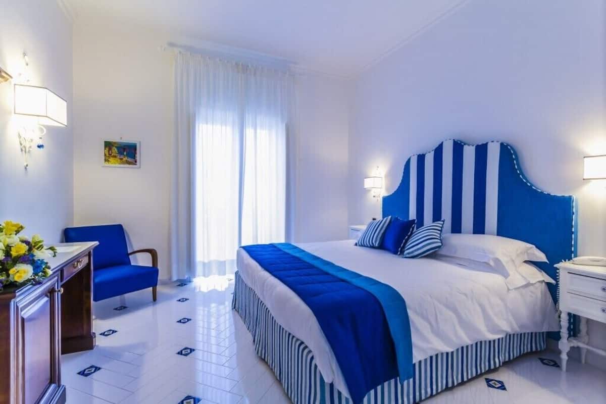 Una habitación de hotel con una cama grande, una silla azul, un escritorio con una lámpara y un cuadro en la pared.