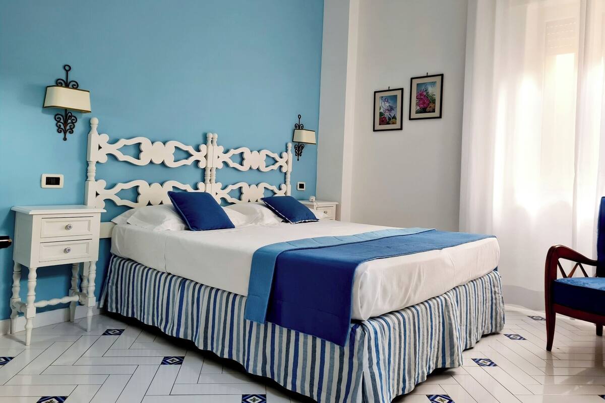 Un dormitorio con cama, mesitas de noche, una silla y una ventana con cortinas.