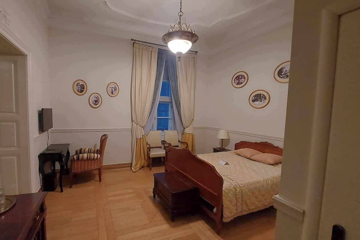 Un dormitori amb un llit, una taula de treball, una cadira, una televisió i una finestra amb cortines.