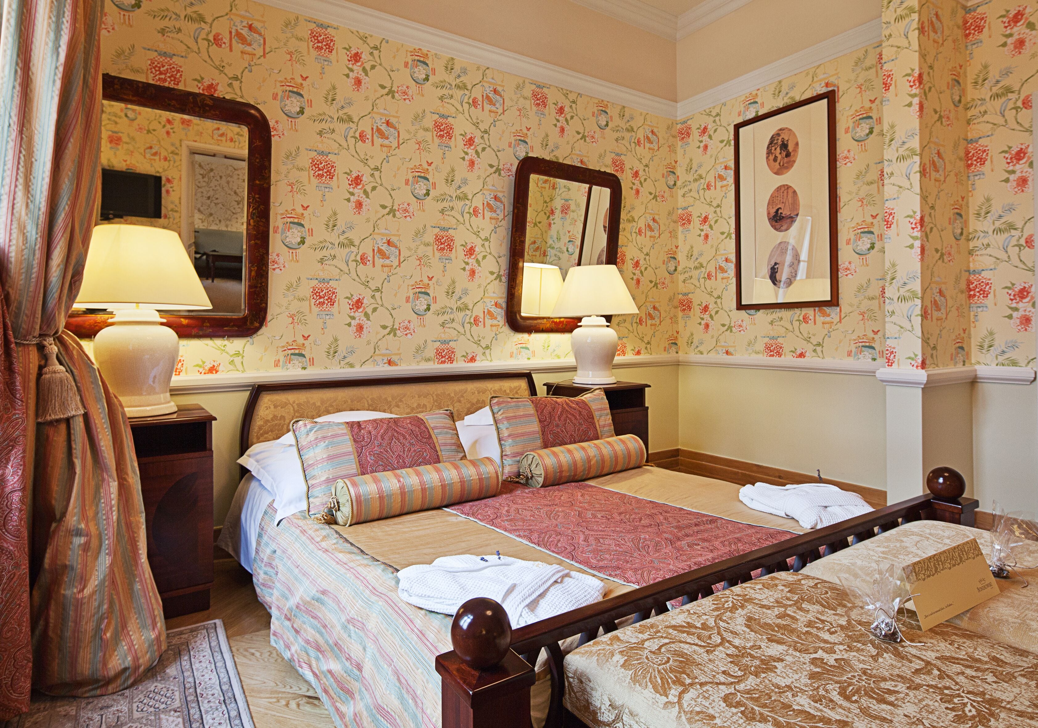 A hotel room with floral wallpaper, a large bed with multiple pillows, two bedside tables with lamps, and a mirror on the wall.