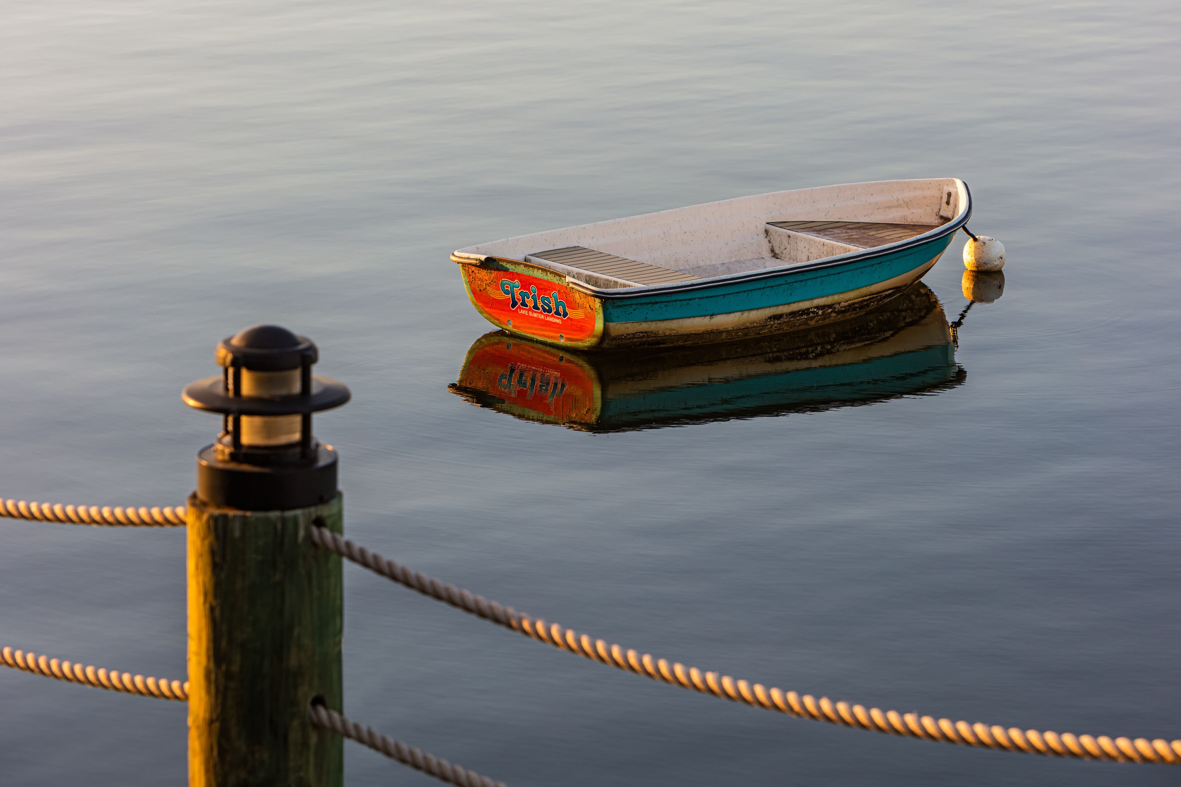 A rowboat named "Trish" tied to a wooden post with ropes.