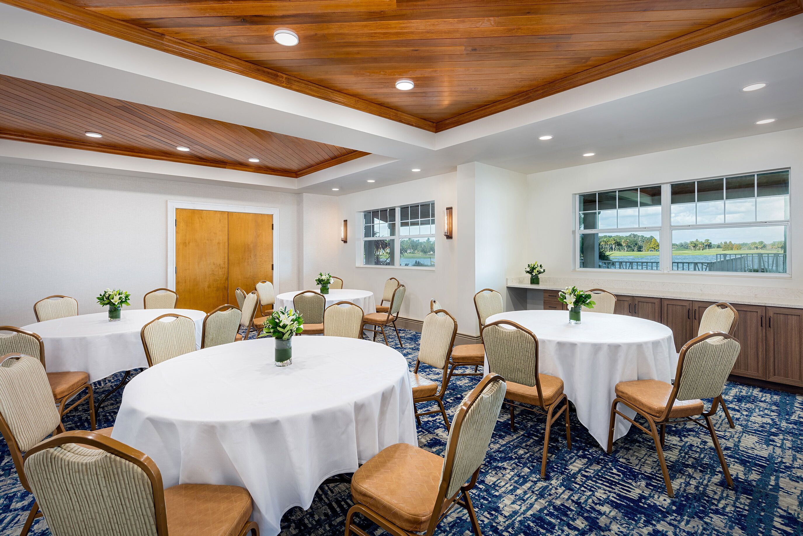 A conference room with round tables, beige chairs, and a view of a lake through large windows.