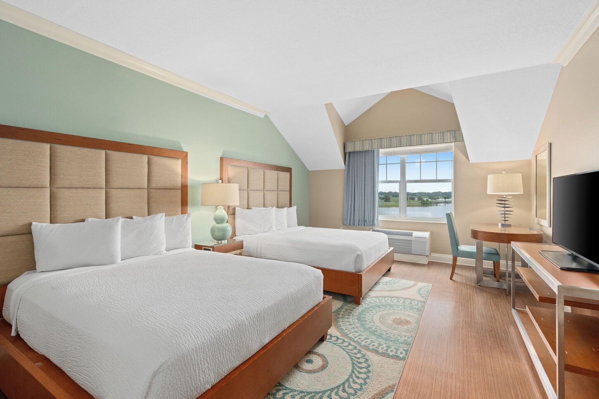A hotel room with two beds, a desk, a TV, and a large window with a view.