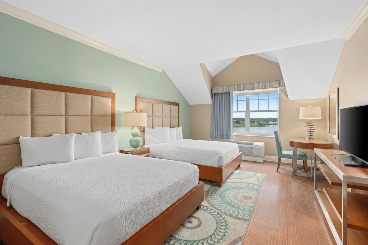 A hotel room with two beds, a desk, a TV, and a large window with a view.