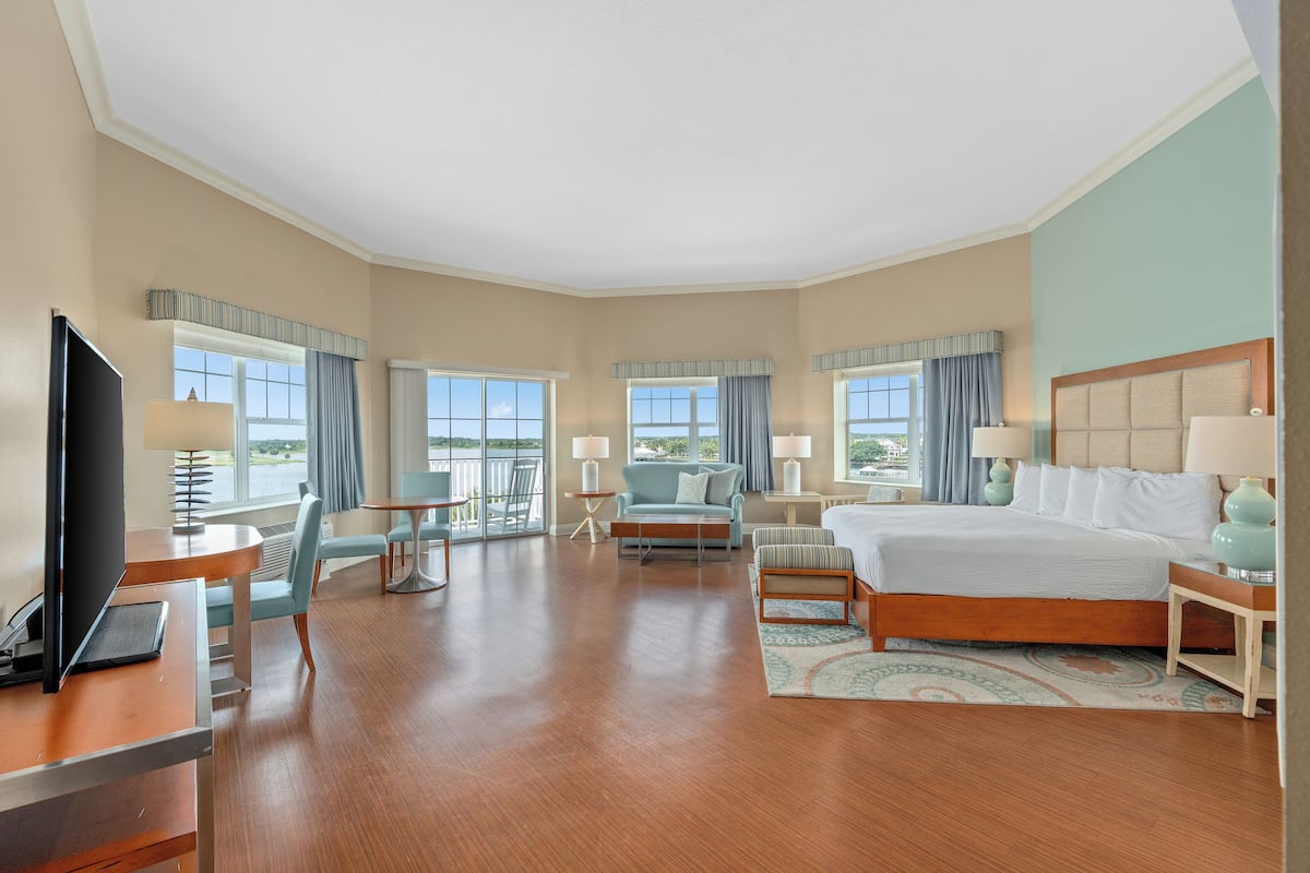 A hotel room with a large bed, a desk with a computer, a chair, a sofa, and a balcony with a view.