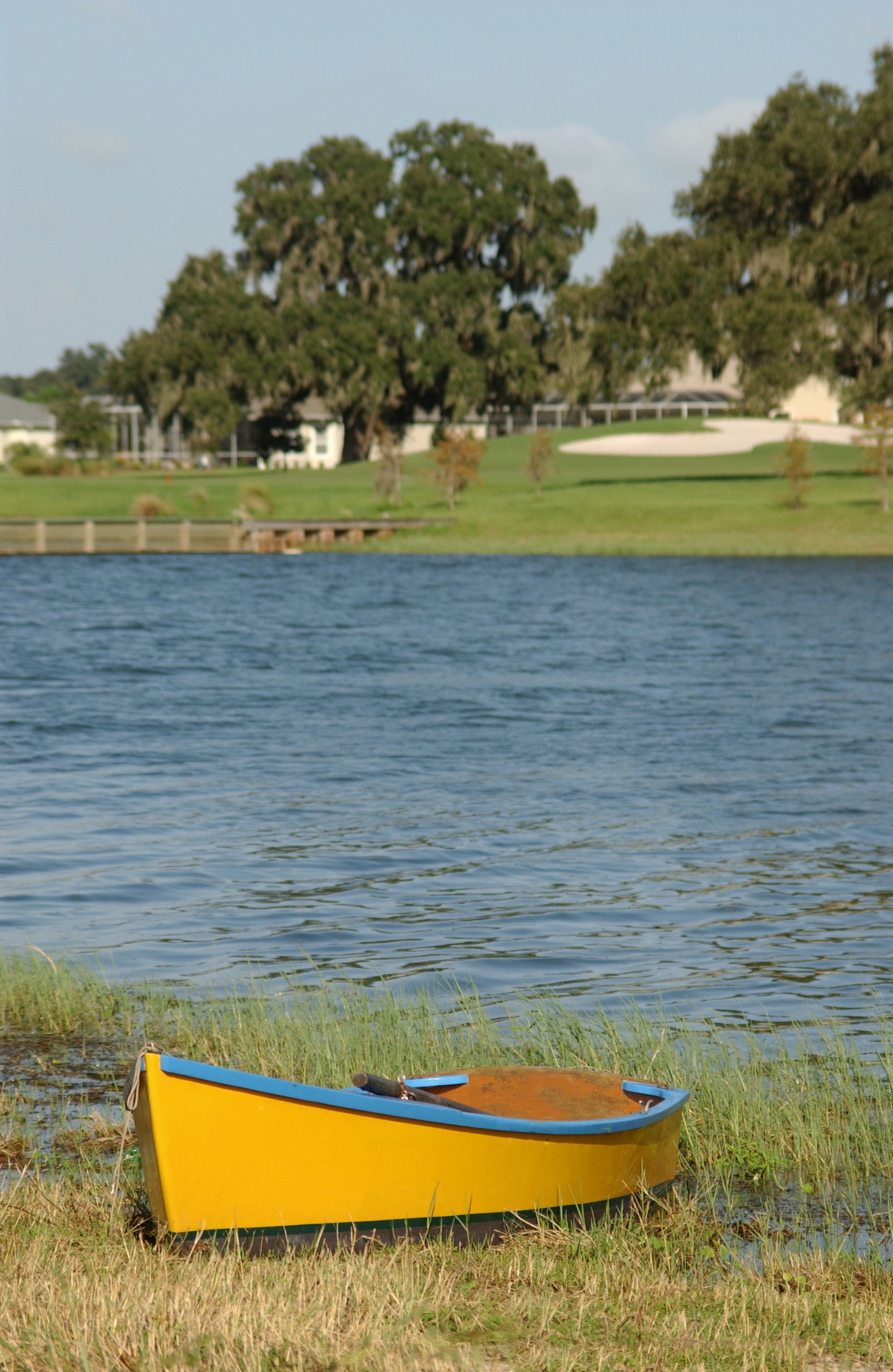 A yellow boat on a grassy shore by a calm lake with trees and houses in the background.