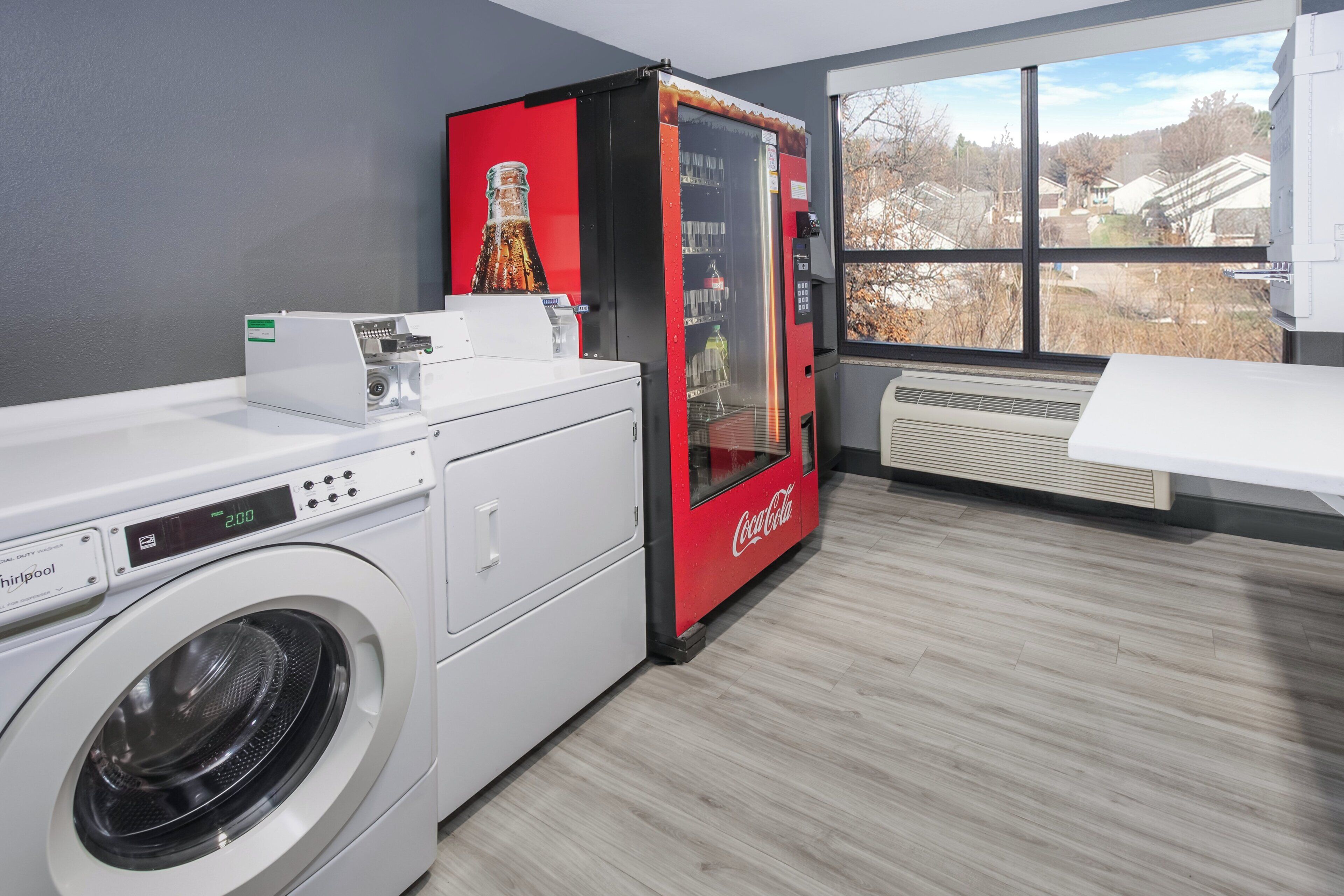 A laundry room with a front-loading washer, a red Coca-Cola refrigerator, and a large window with a view of houses.