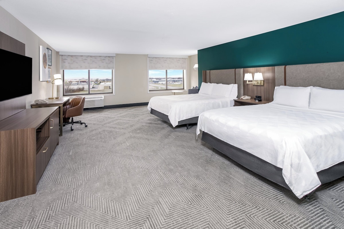 A hotel room with two beds, a desk, a chair, a TV, and large windows with a view.