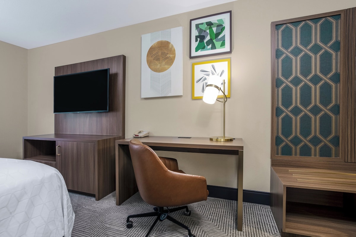 A hotel room with a bed, a desk with a chair, a television, and artwork on the wall.