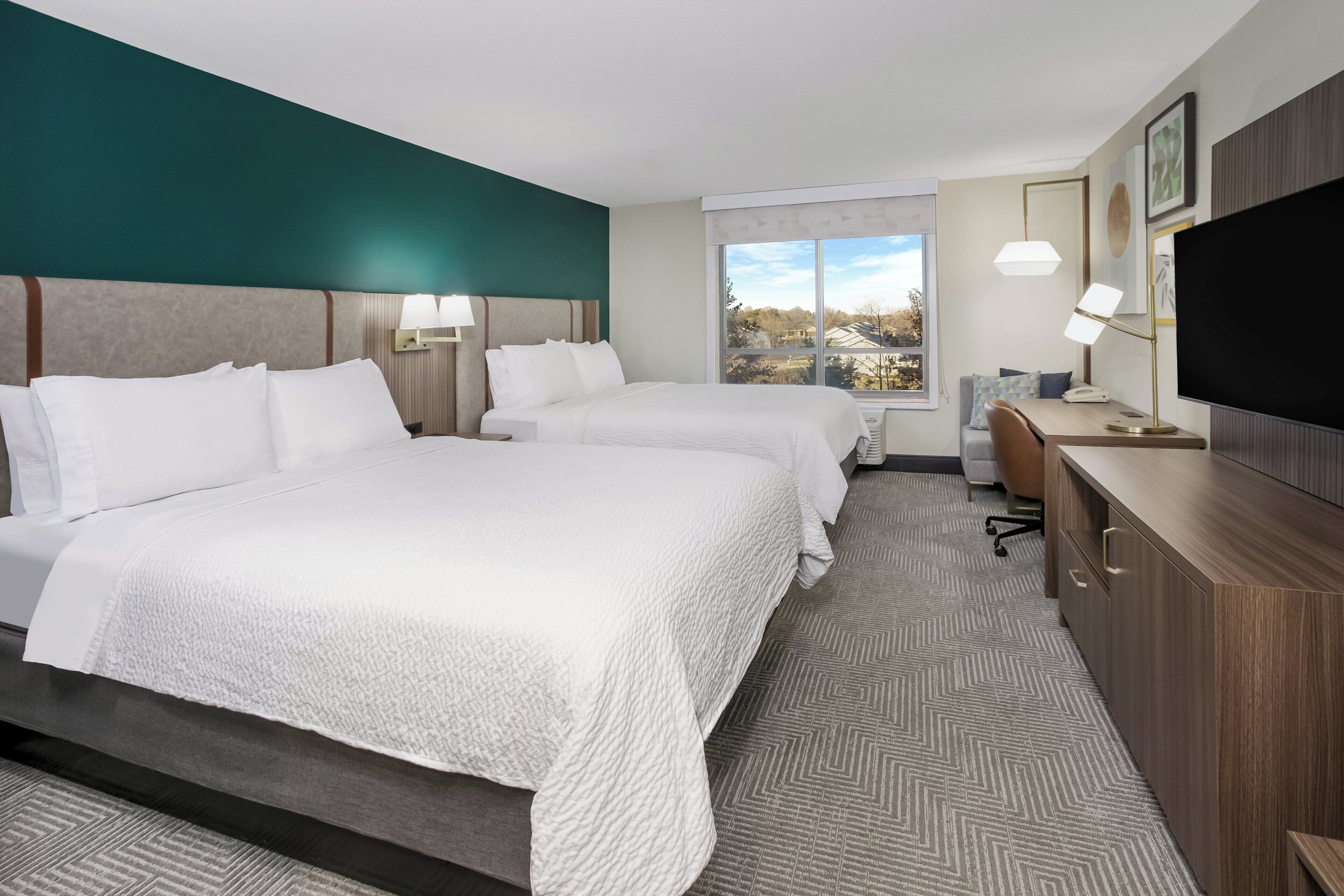 A hotel room with two beds, a desk, a TV, and a window with a view.