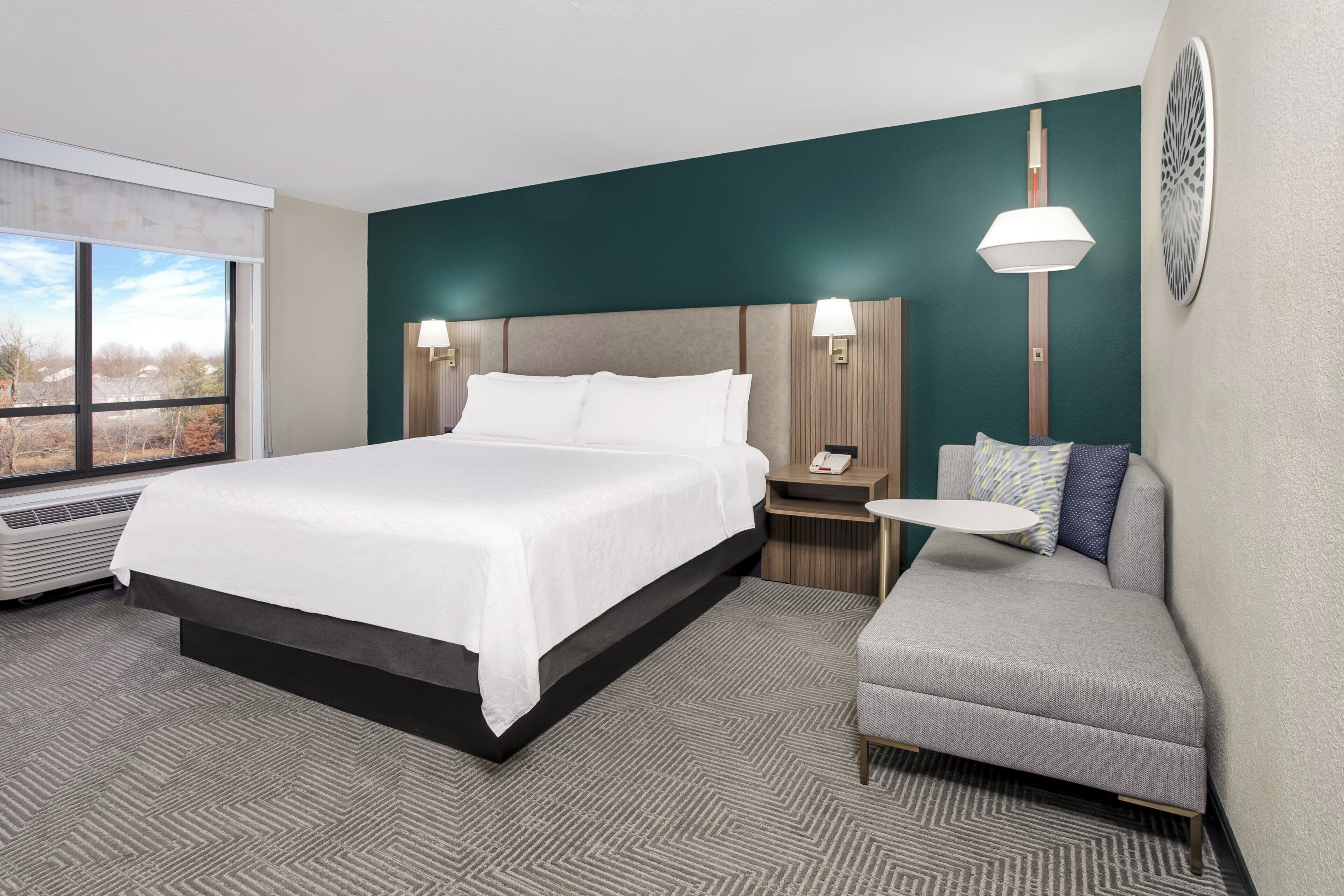 Habitación de hotel con una cama grande, un sofá pequeño, una mesa redonda y una lámpara de pared.