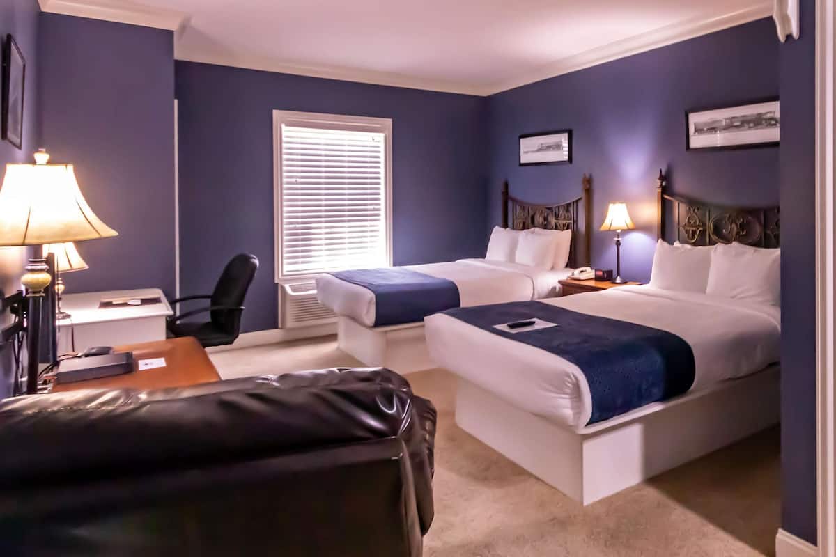 A hotel room with two beds, a desk, a chair, a lamp, a window with blinds, a bathroom with a toilet and towels, and a picture on the wall.