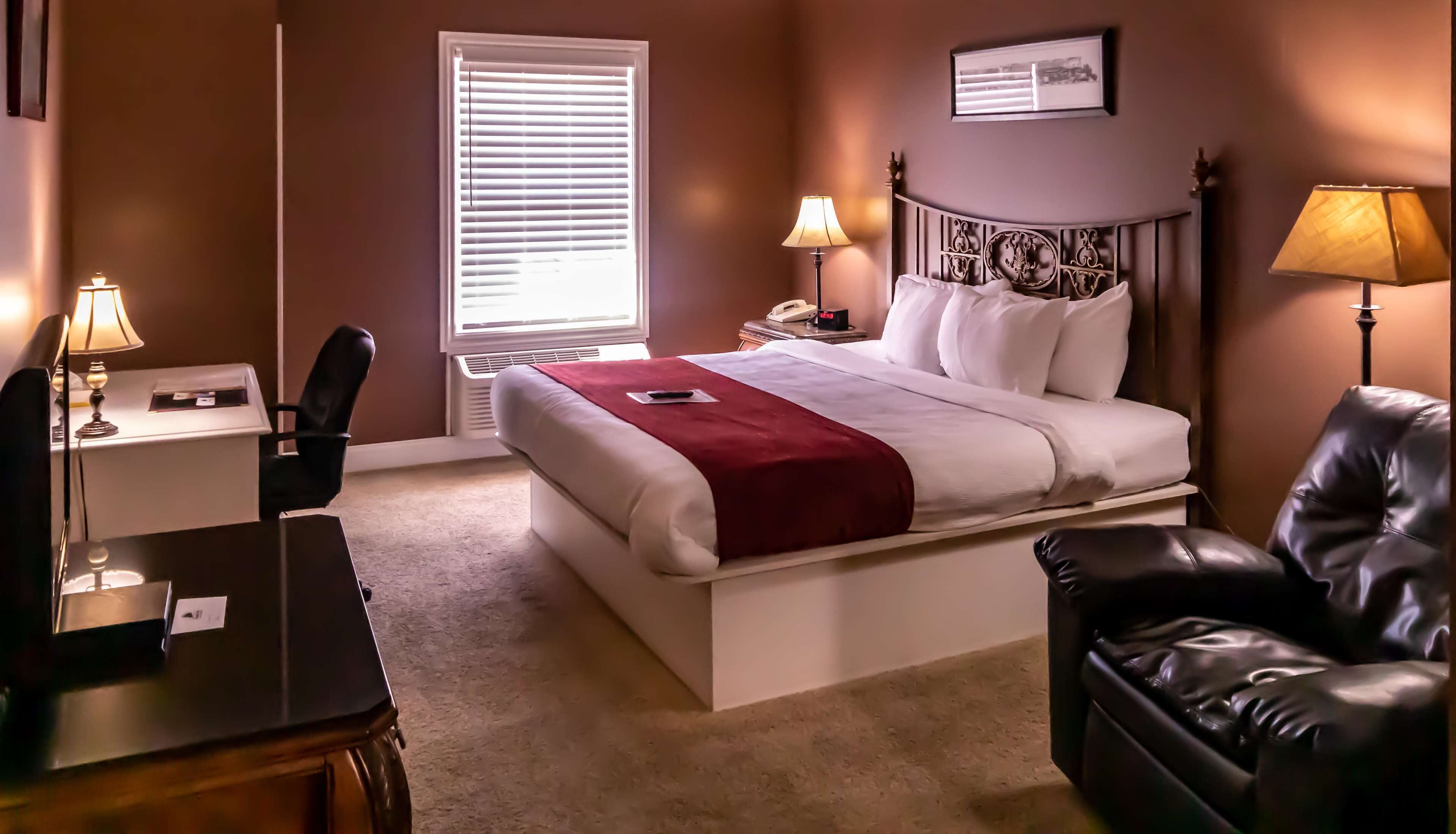 A hotel room with a bed, two bedside lamps, a desk, a chair, and a leather armchair.