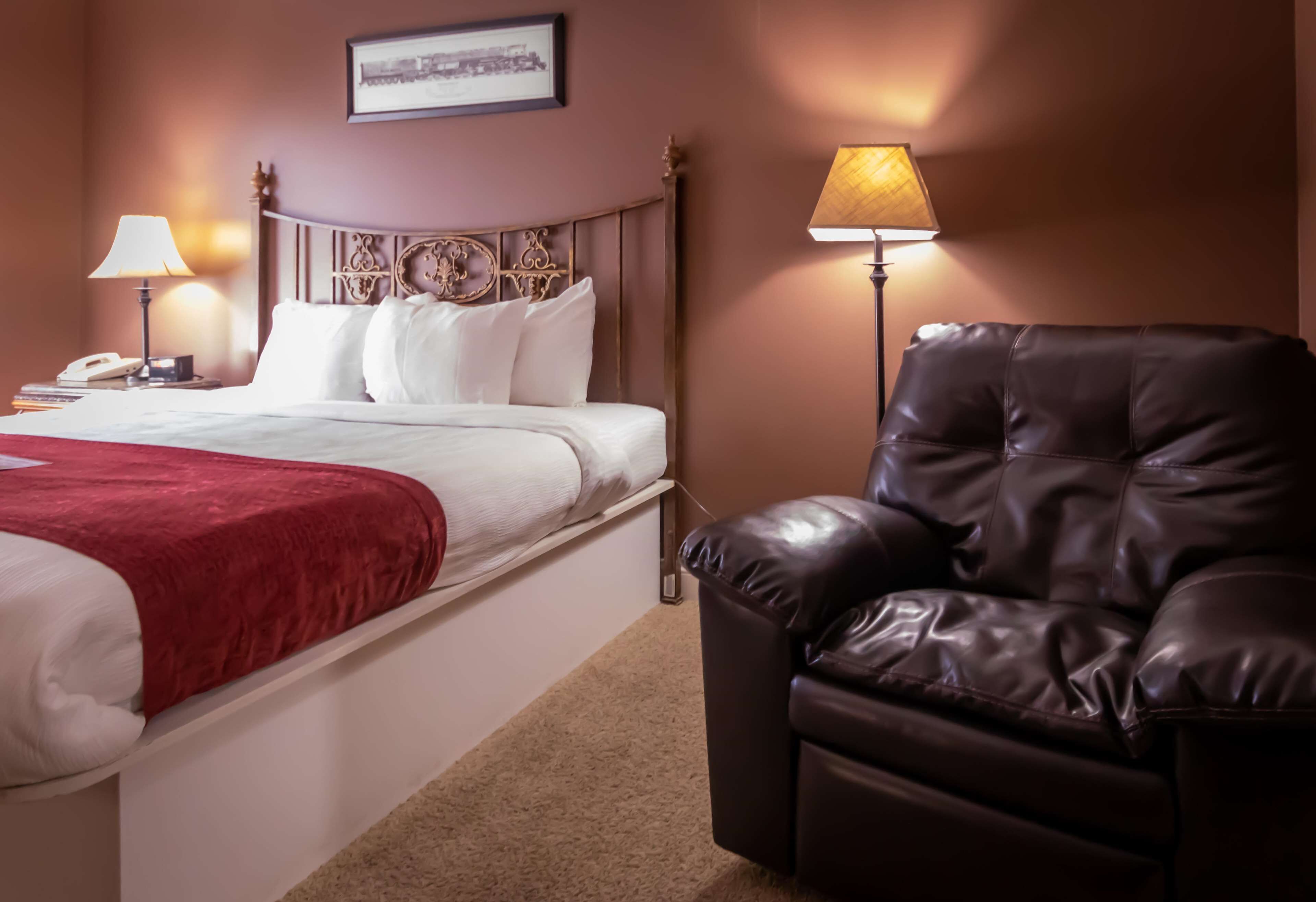 A hotel room with a bed, a leather armchair, two bedside lamps, a nightstand with a phone, and a framed picture on the wall.