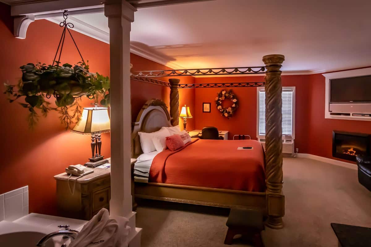 A four-poster bed with a red bedspread, a bedside table with a lamp, a television mounted on the wall, and a fireplace.