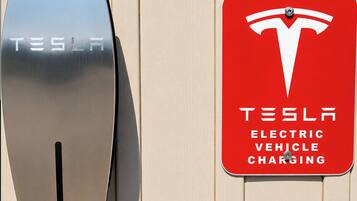A Tesla electric vehicle charging station with a metallic charging unit and a red sign indicating the service.
