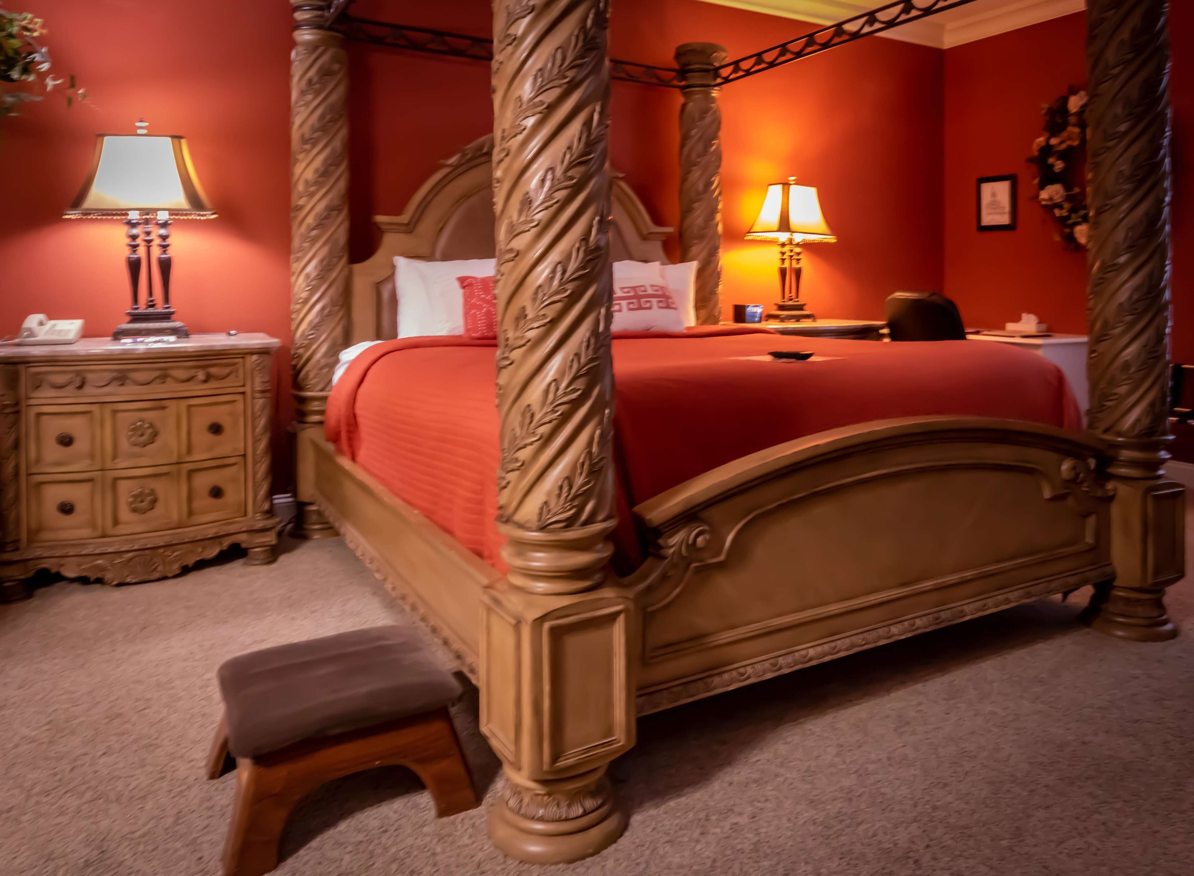 Een hemelbed met een rode sprei, een houten nachtkastje met een lamp en een klein bankje.