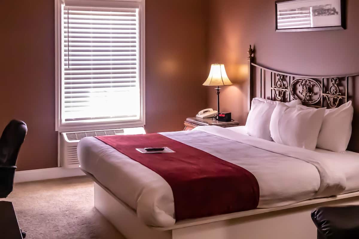 A hotel room with a large bed, a desk with a lamp, a telephone, and a framed picture on the wall.