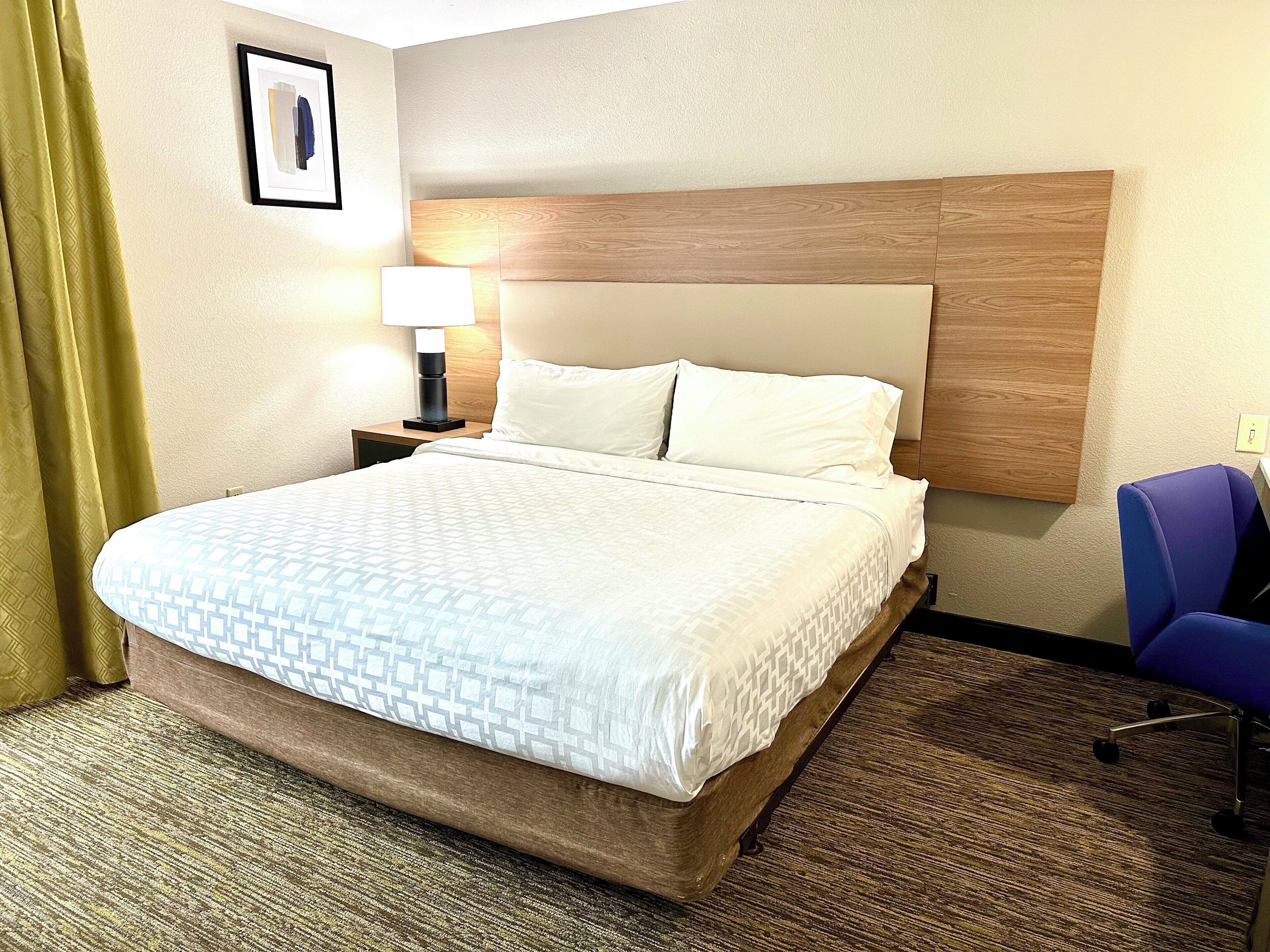 A hotel room with a large bed, a nightstand with a lamp, a blue chair, and a framed picture on the wall.