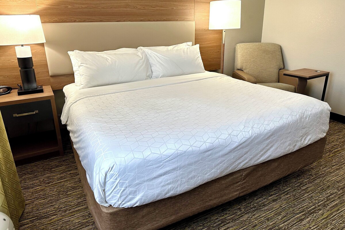 Ein Hotelzimmer mit einem großen Bett, zwei Nachttischlampen, einem Sessel, einem kleinen Tisch und einem Nachttisch mit Lampe und Telefon.