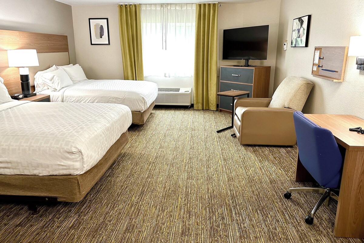 Ein Hotelzimmer mit zwei Betten, einem Schreibtisch, einem Stuhl, einem Fernseher und einem Fenster mit Vorhängen.