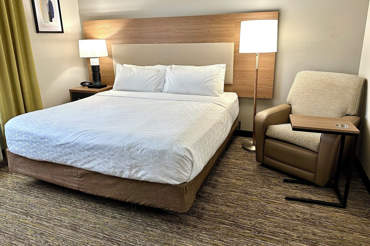 Ein Hotelzimmer mit einem großen Bett, zwei Nachttischlampen, einem Sessel mit Tablett und einem gerahmten Bild an der Wand.